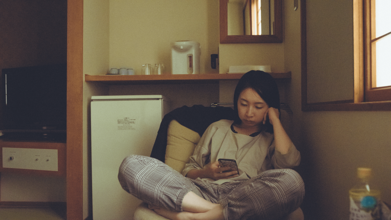Young woman sitting in her kitchen and looking at her phone, representing everyday scrolling and the theme of doomscrolling.