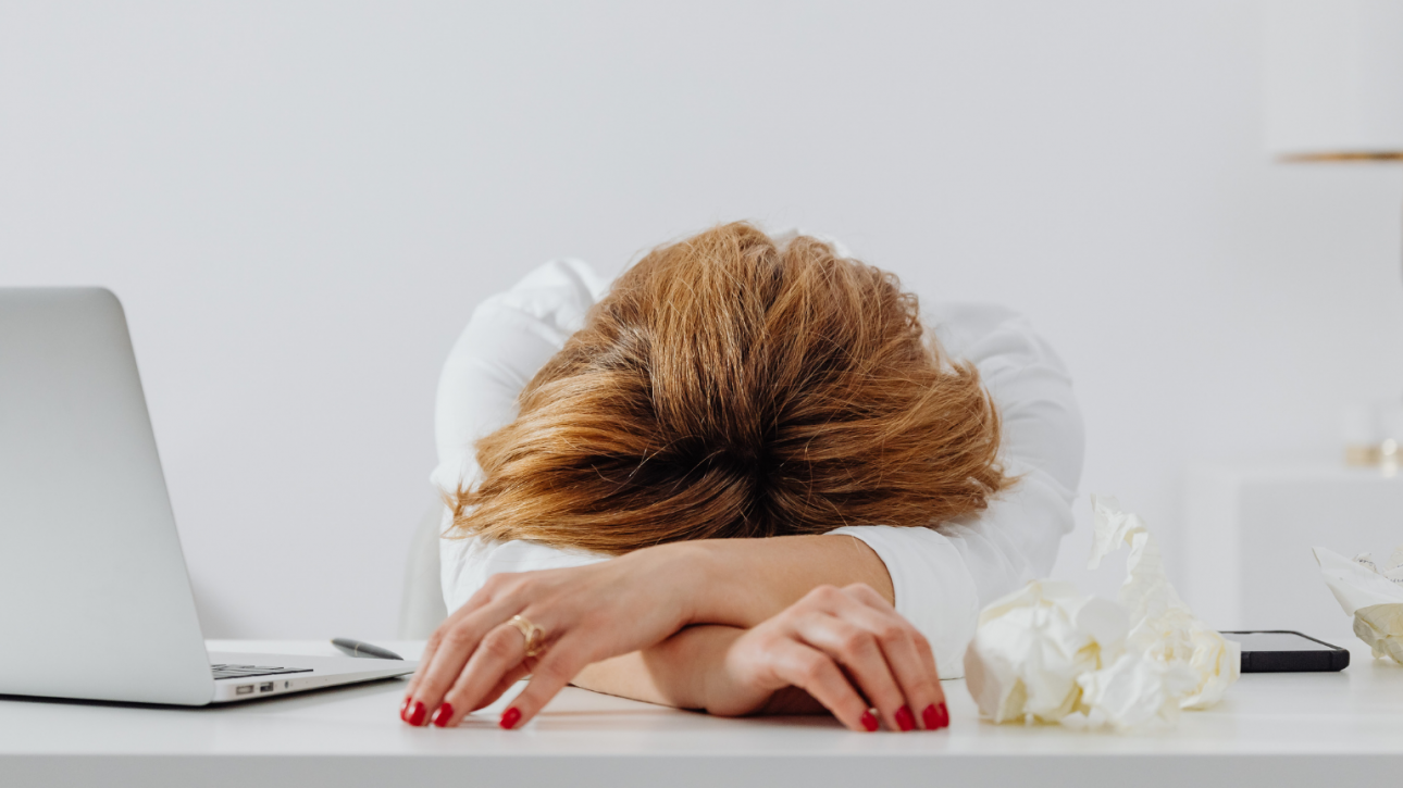 A professional woman resting her head on her desk, appearing exhausted, symbolising the emotional toll of perfectionism.
