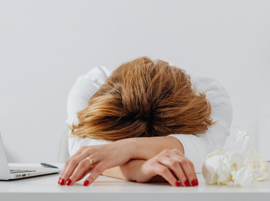 A professional woman resting her head on her desk, appearing exhausted, symbolising the emotional toll of perfectionism.