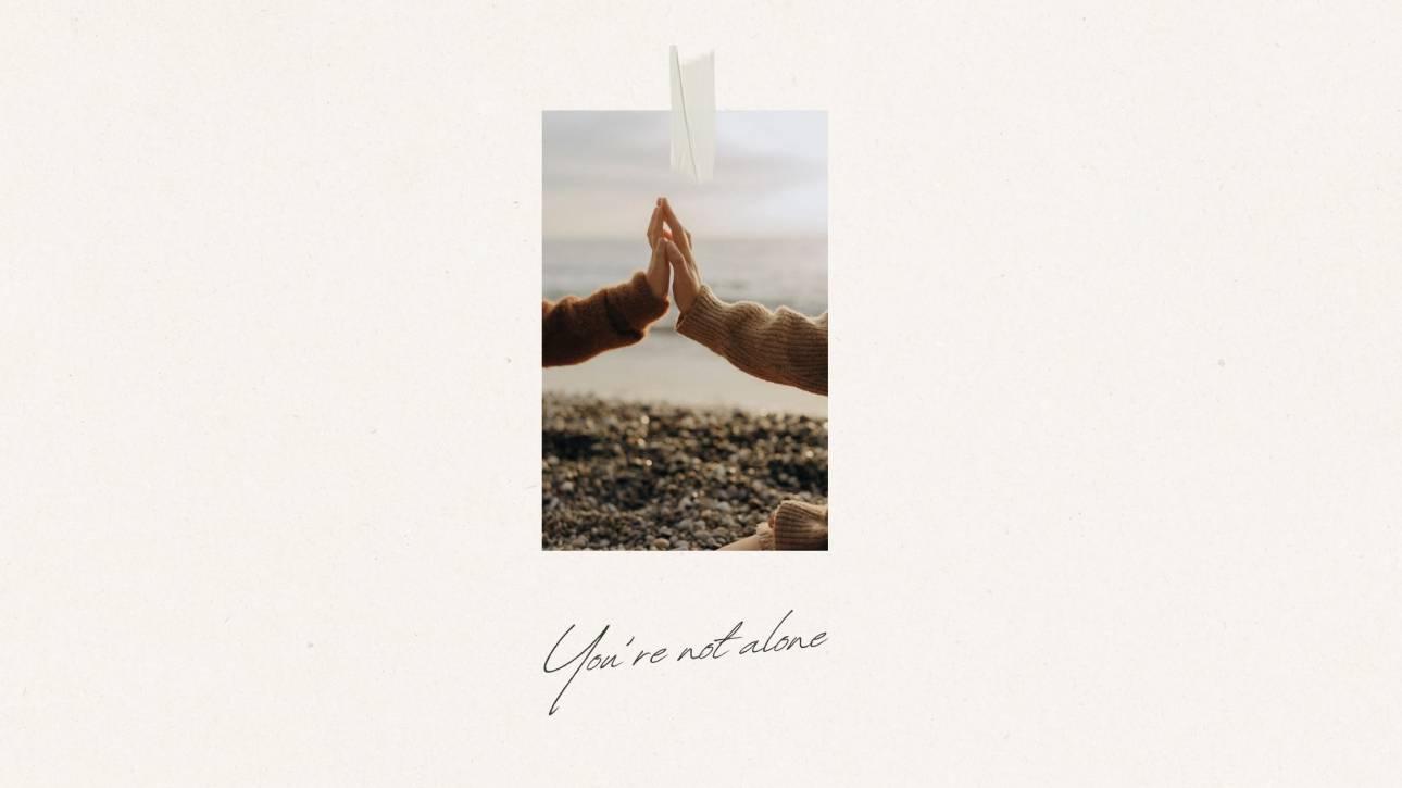 Two people in cosy knitwear touching hands on a pebble beach, representing connection and community in eating disorder recovery