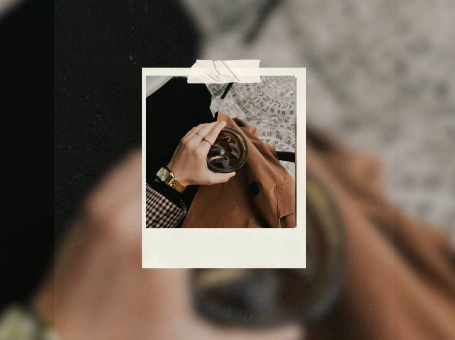 Aerial view of woman's hand holding iced coffee, wearing tweed jacket and gold jewelry, representing taking yourself on a date