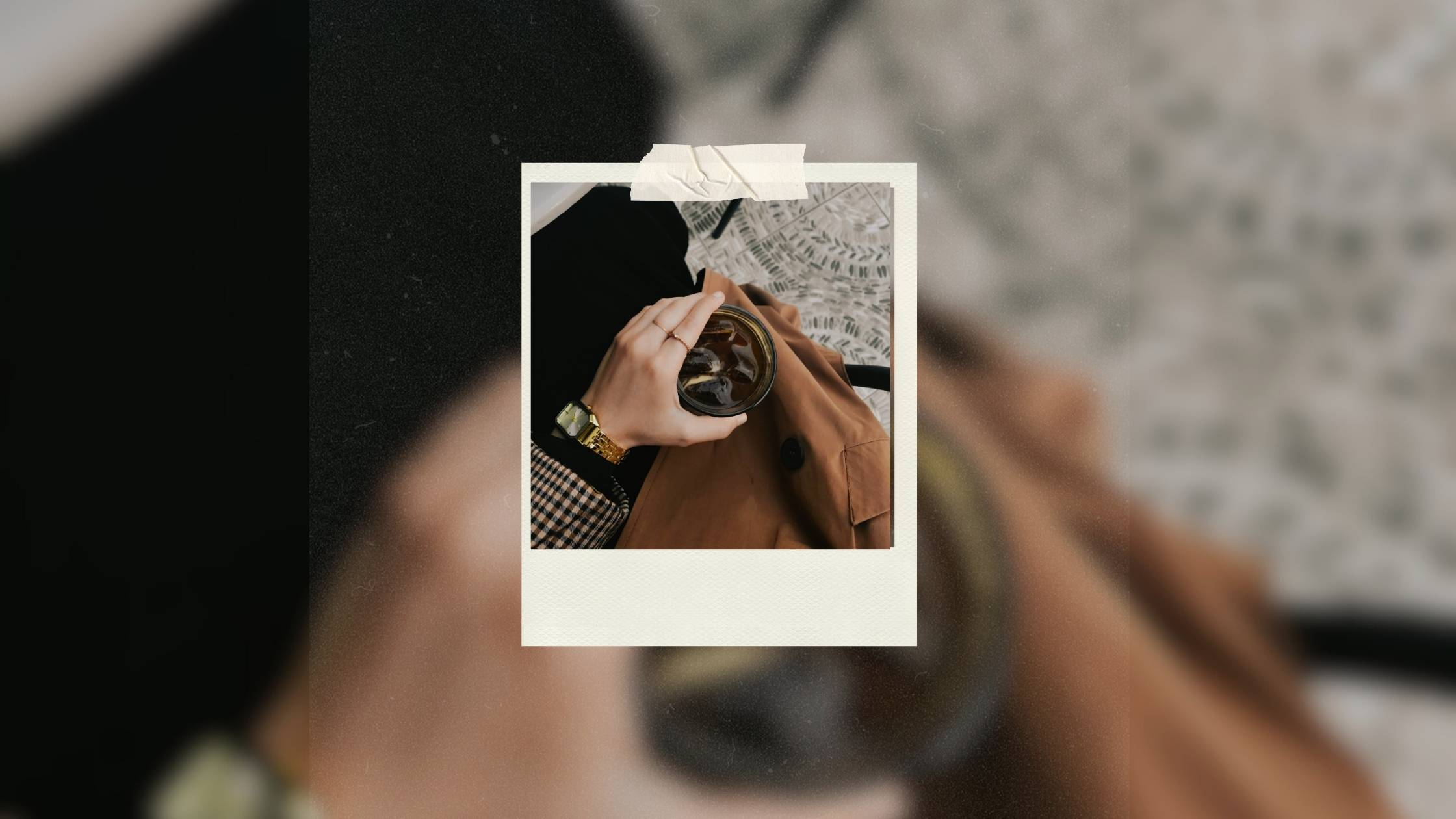 Aerial view of woman's hand holding iced coffee, wearing tweed jacket and gold jewelry, representing taking yourself on a date