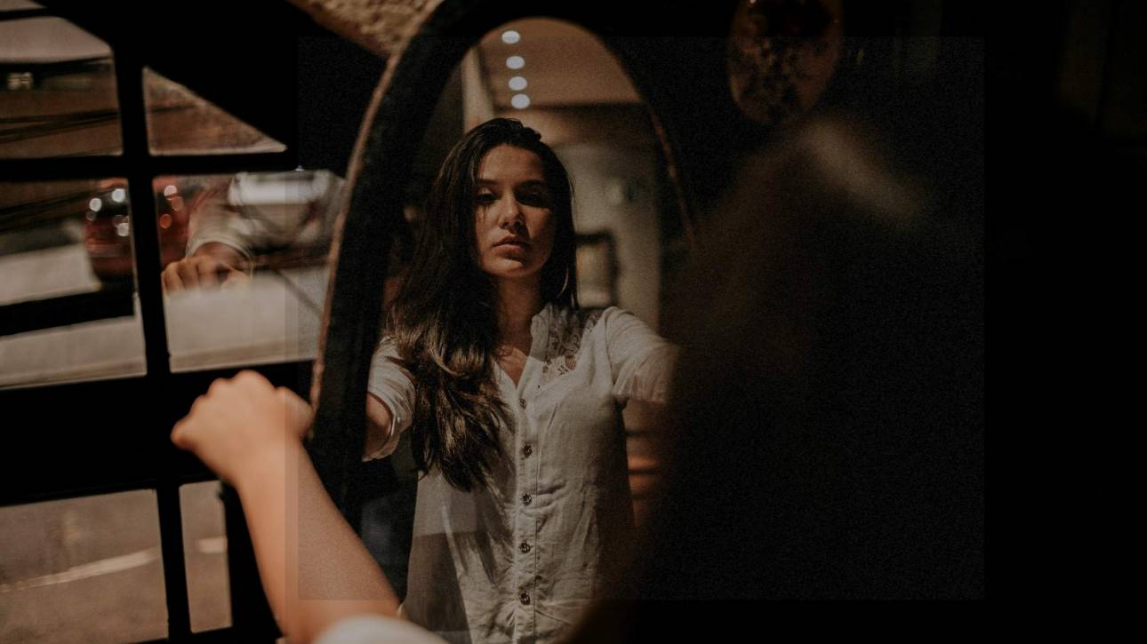 Young woman studying her reflection in an old-fashioned oval full-length mirror, her actual body blurred while reflection is in focus, representing self-examination and inner dialogue
