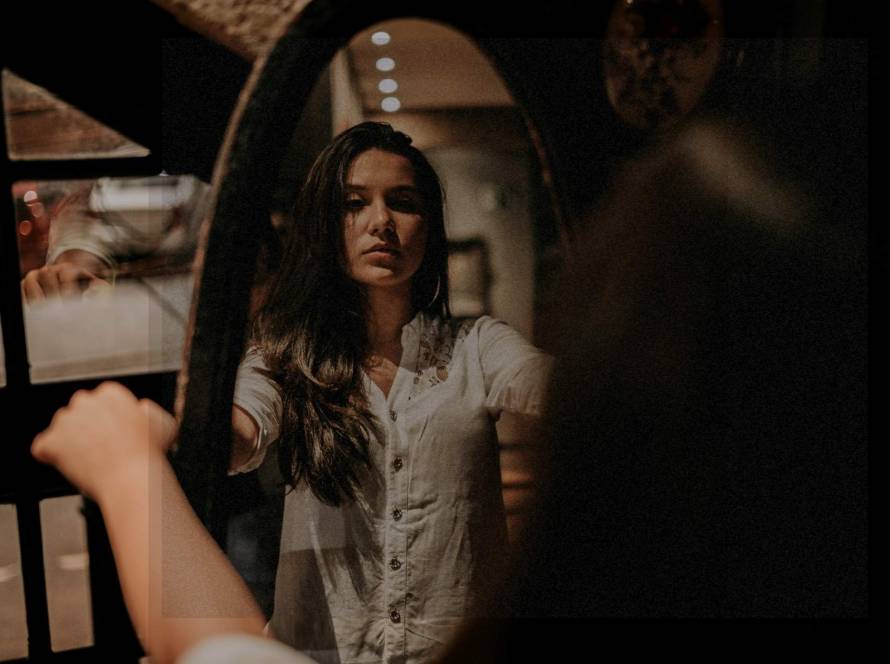 Young woman studying her reflection in an old-fashioned oval full-length mirror, her actual body blurred while reflection is in focus, representing self-examination and inner dialogue