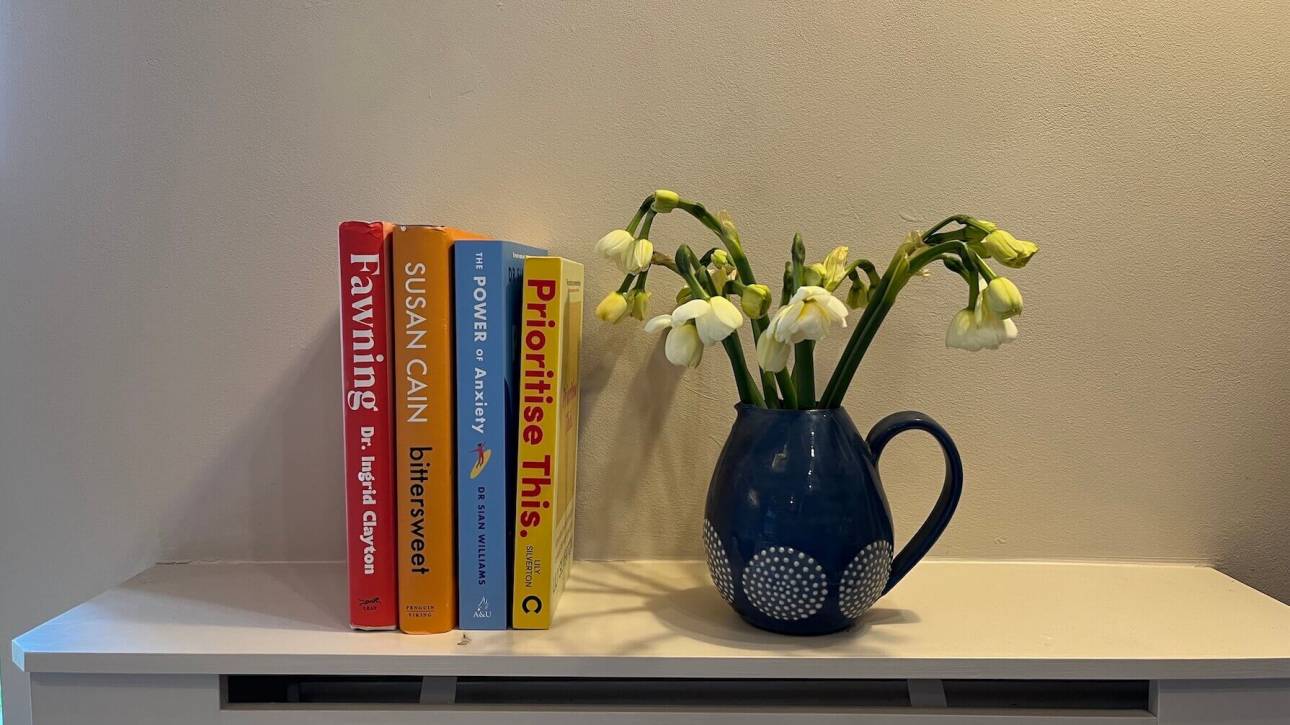 Four books on a light coloured surface beside a blue jug of pale yellow spring flowers — Fawning by Dr Ingrid Clayton, Bittersweet by Susan Cain, The Power of Anxiety by Dr Sian Williams, and Prioritise This by Lily Silverton