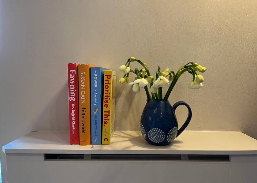 Four books on a light coloured surface beside a blue jug of pale yellow spring flowers — Fawning by Dr Ingrid Clayton, Bittersweet by Susan Cain, The Power of Anxiety by Dr Sian Williams, and Prioritise This by Lily Silverton