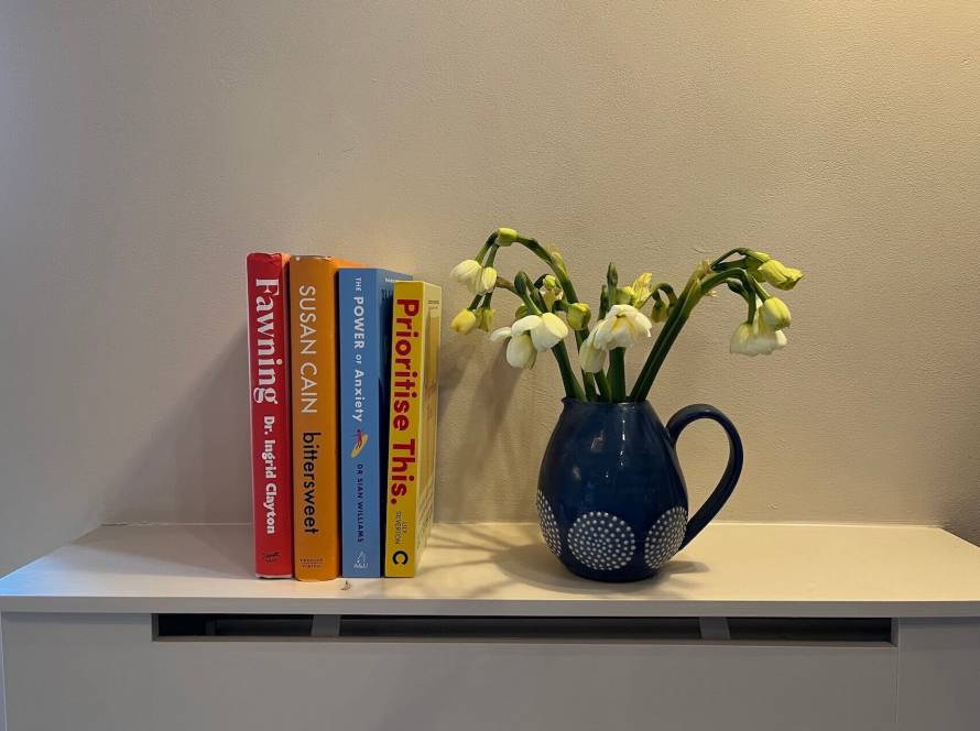 Four books on a light coloured surface beside a blue jug of pale yellow spring flowers — Fawning by Dr Ingrid Clayton, Bittersweet by Susan Cain, The Power of Anxiety by Dr Sian Williams, and Prioritise This by Lily Silverton