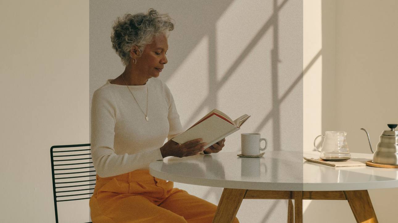 A woman over 50 sitting quietly at a table with a book and coffee, representing the space therapy can offer during midlife transitions