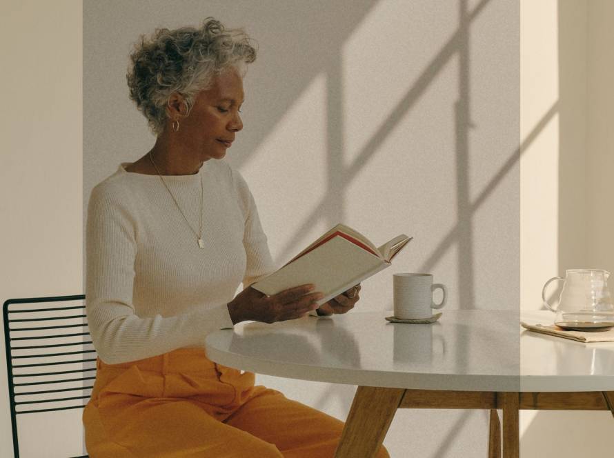 A woman over 50 sitting quietly at a table with a book and coffee, representing the space therapy can offer during midlife transitions