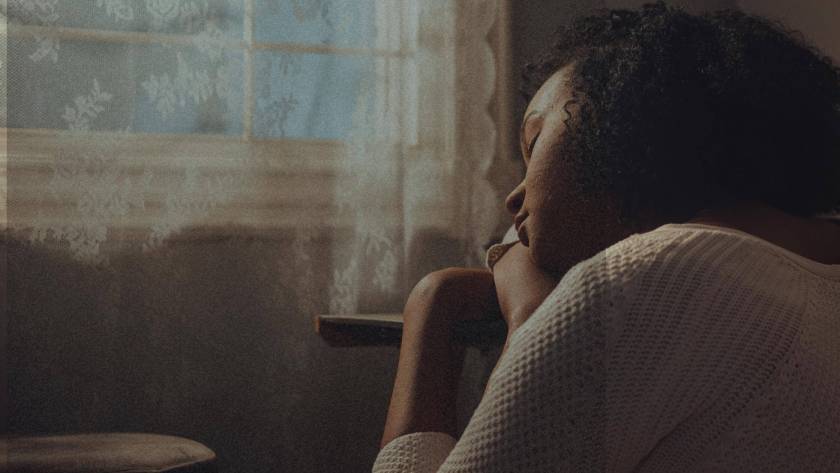 A woman sitting in a chair in quiet contemplation, photographed from behind in warm, atmospheric light with a lace curtain in the background.