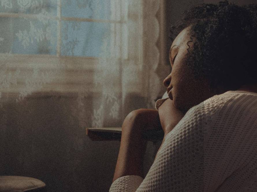 A woman sitting in a chair in quiet contemplation, photographed from behind in warm, atmospheric light with a lace curtain in the background.