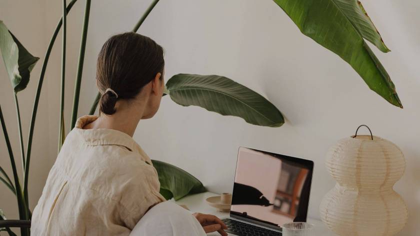 Woman seen from behind sitting at a desk working on a laptop, surrounded by large tropical plants in warm neutral tones, composed and unreachable