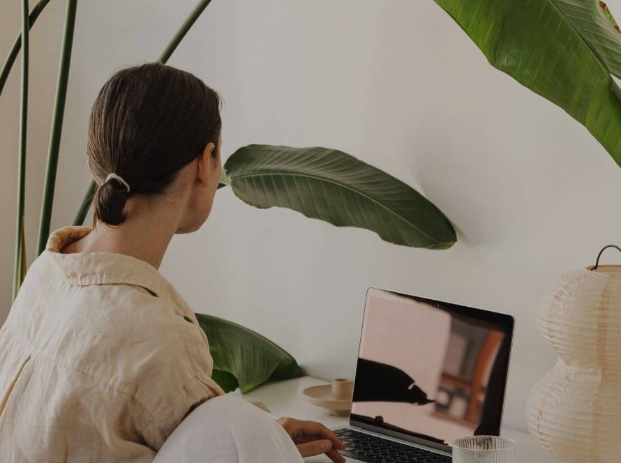 Woman seen from behind sitting at a desk working on a laptop, surrounded by large tropical plants in warm neutral tones, composed and unreachable