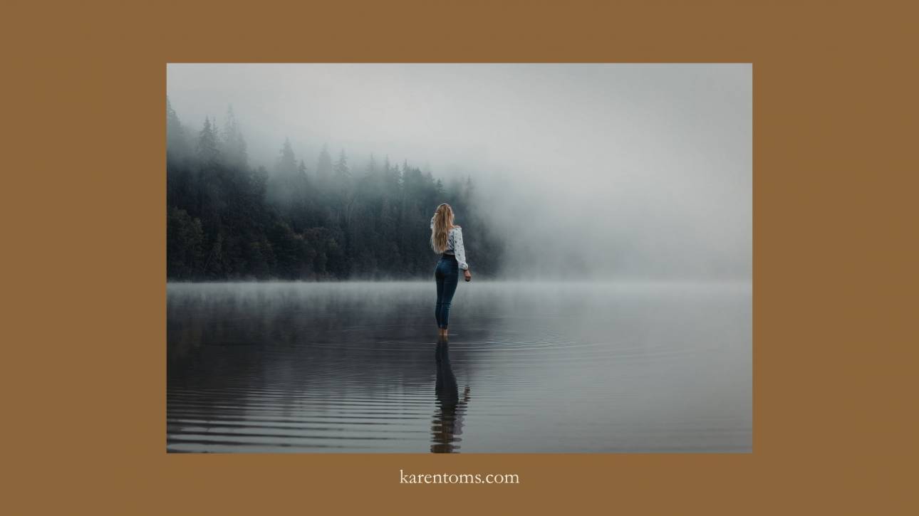Woman standing alone in a misty lake from behind, surrounded by fog and dark trees, figure small within a vast atmospheric landscape