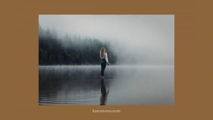 Woman standing alone in a misty lake from behind, surrounded by fog and dark trees, figure small within a vast atmospheric landscape
