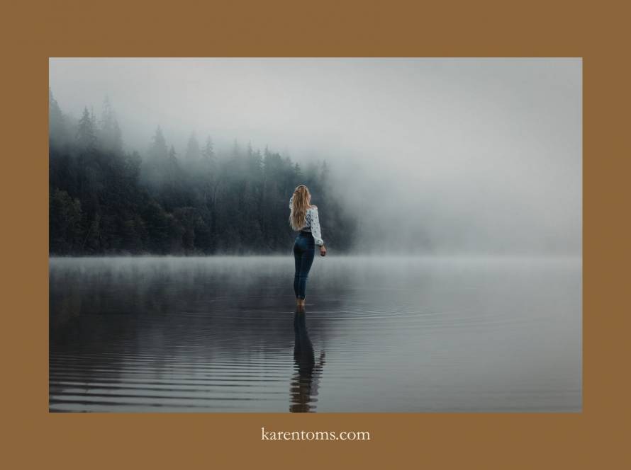Woman standing alone in a misty lake from behind, surrounded by fog and dark trees, figure small within a vast atmospheric landscape