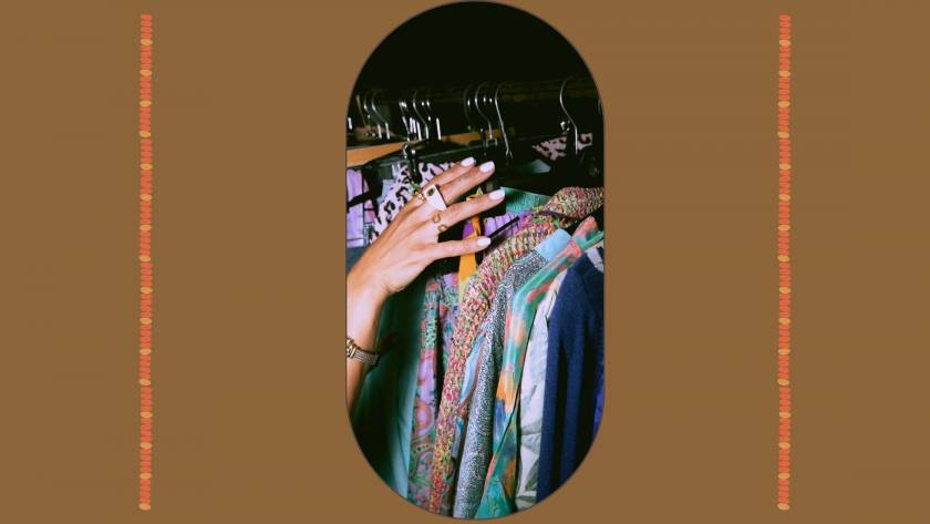 A hand with gold rings reaching toward a rail of patterned silk and printed clothing in jewel tones, against a warm brown background, illustrating the quiet act of choosing clothes that express who you are.
