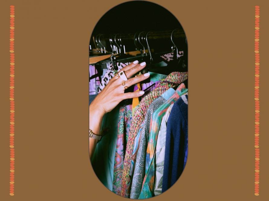A hand with gold rings reaching toward a rail of patterned silk and printed clothing in jewel tones, against a warm brown background, illustrating the quiet act of choosing clothes that express who you are.