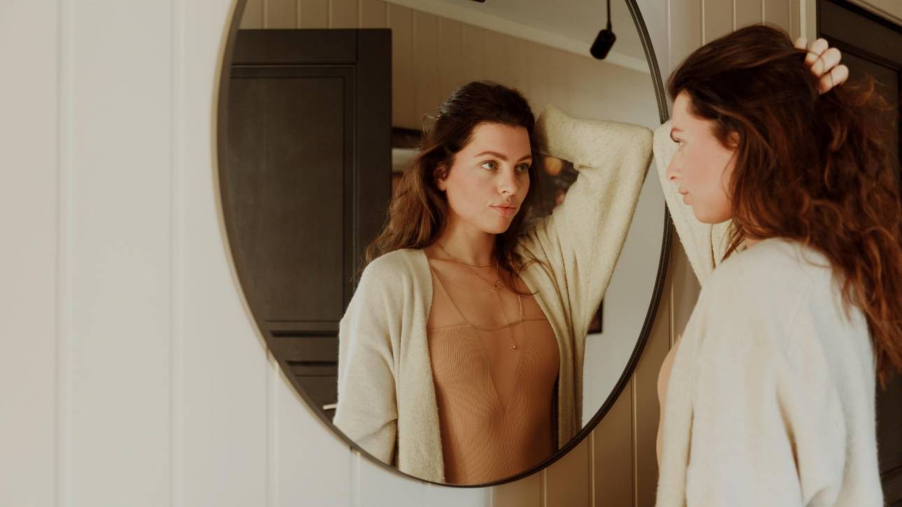 A woman in a cream cardigan and tan top reflected in a round mirror, appraising herself, illustrating the inner critic at work in the moment of getting dressed.