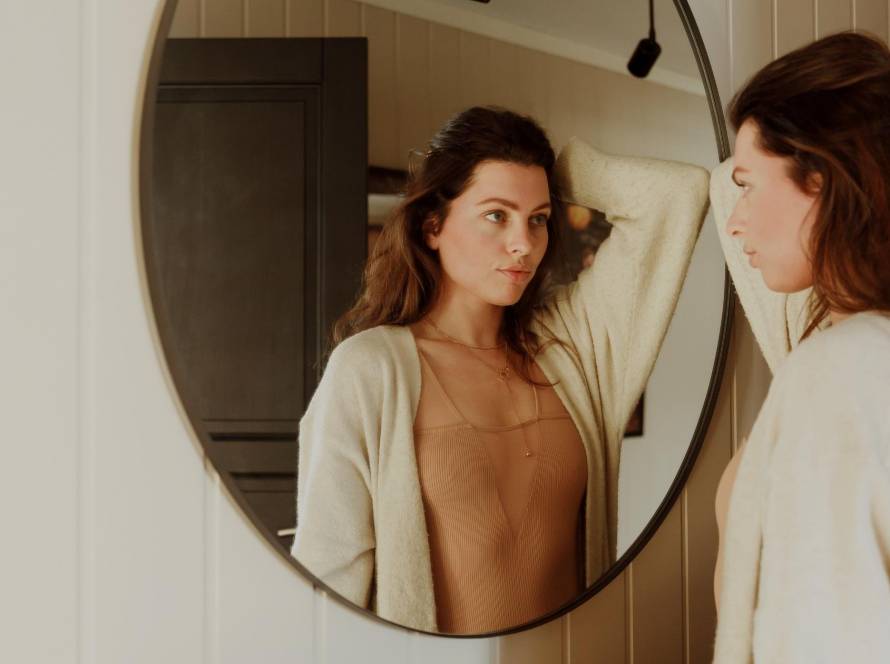 A woman in a cream cardigan and tan top reflected in a round mirror, appraising herself, illustrating the inner critic at work in the moment of getting dressed.