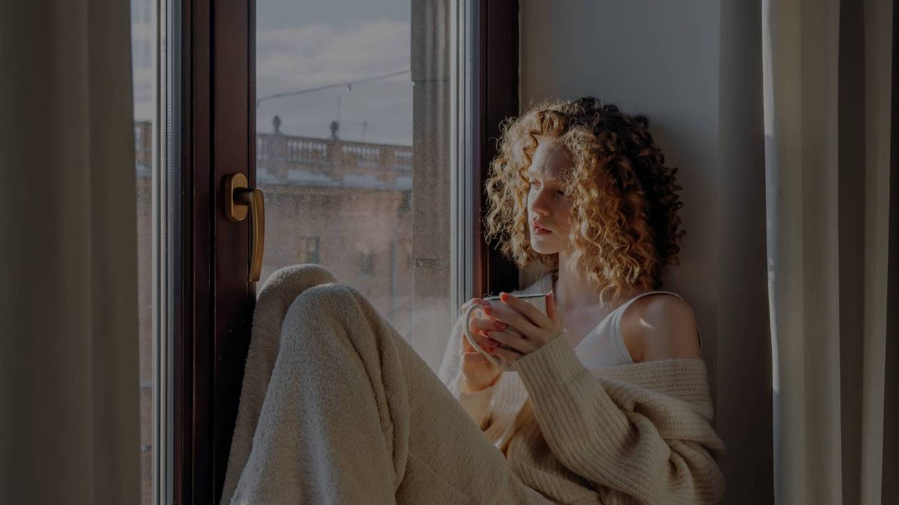 A woman in a cream knitted cardigan and white socks sitting on a bed in a window seat, holding a small mug, with warm natural light coming through the window — illustrating the quiet pause of burnout recovery.