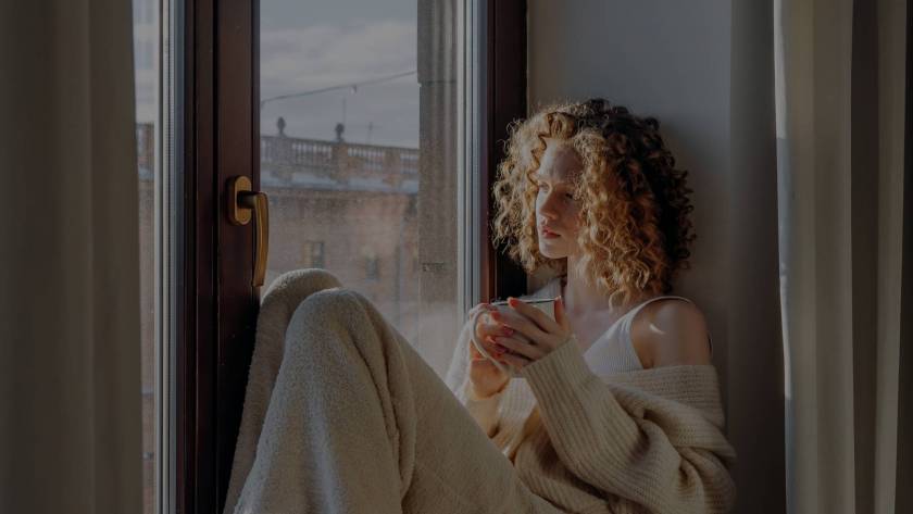 A woman in a cream knitted cardigan and white socks sitting on a bed in a window seat, holding a small mug, with warm natural light coming through the window — illustrating the quiet pause of burnout recovery.