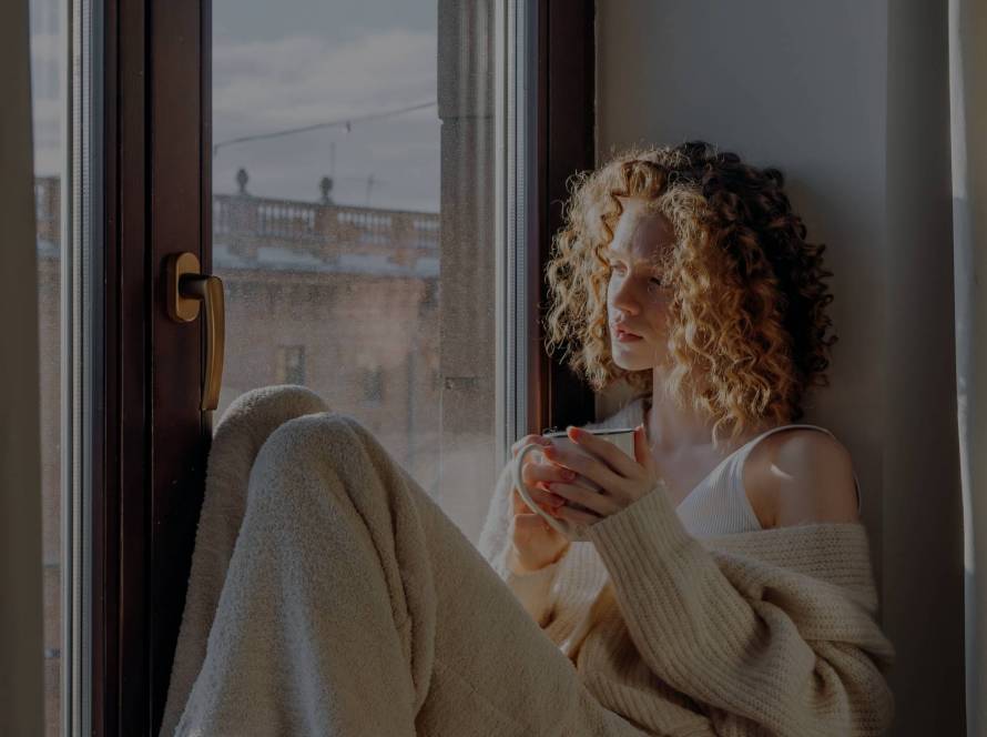 A woman in a cream knitted cardigan and white socks sitting on a bed in a window seat, holding a small mug, with warm natural light coming through the window — illustrating the quiet pause of burnout recovery.