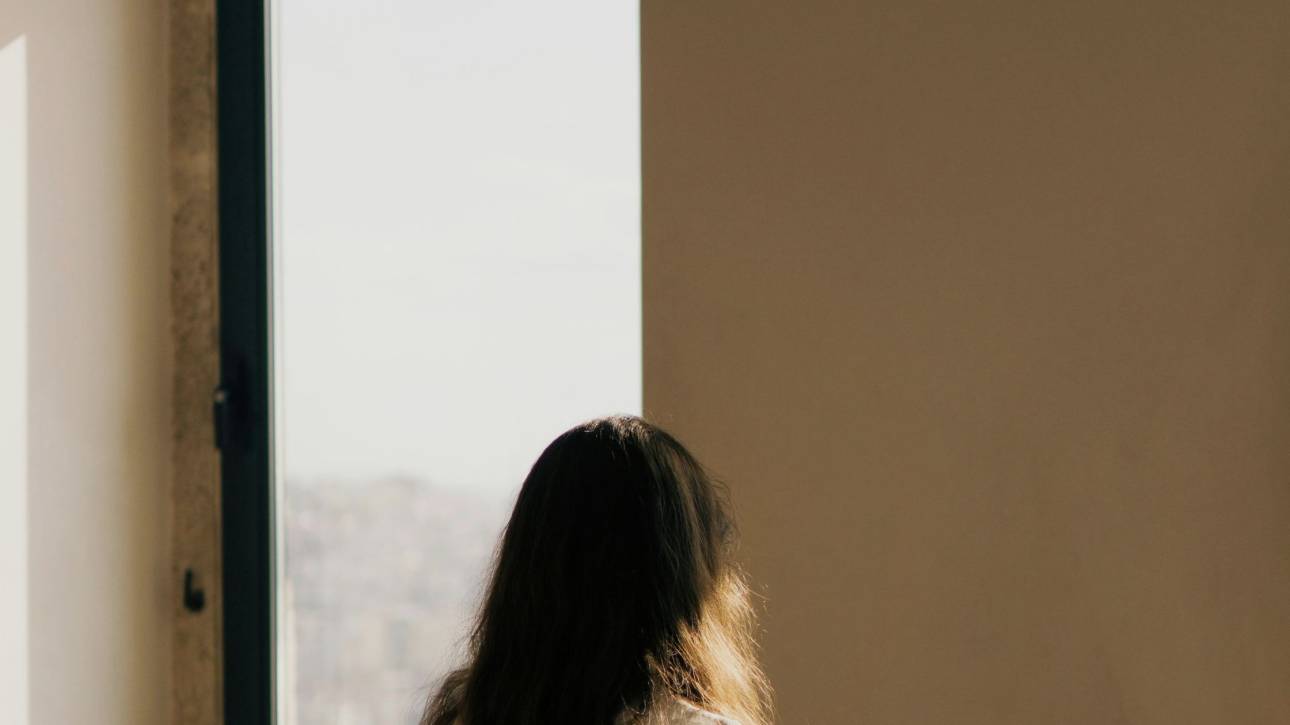 A figure standing small within a large architectural space by an arched window in warm light, illustrating a post about the shy child and self-perception