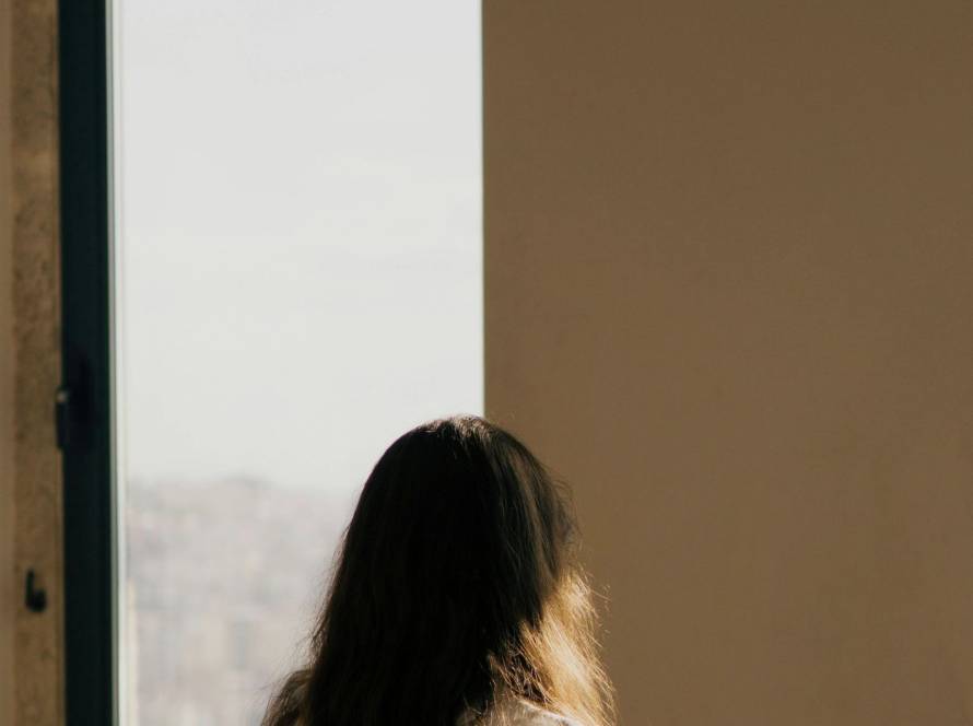 A figure standing small within a large architectural space by an arched window in warm light, illustrating a post about the shy child and self-perception