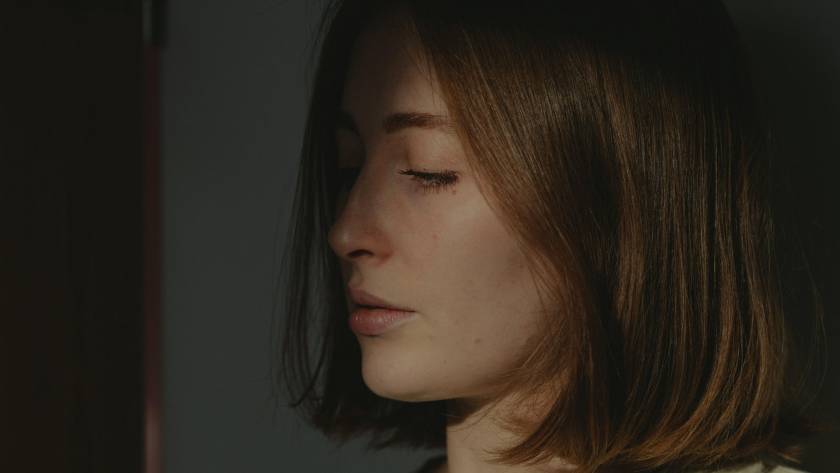Woman in profile with eyes closed, soft directional light against a shadowed wall, reflecting the moment of holding back a known answer