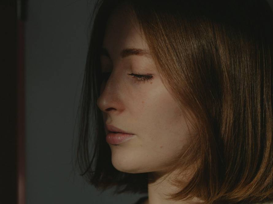 Woman in profile with eyes closed, soft directional light against a shadowed wall, reflecting the moment of holding back a known answer