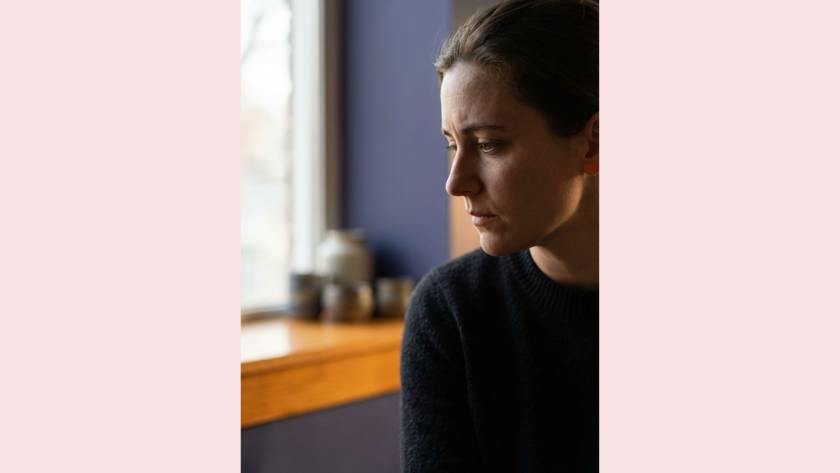 Woman looking away in soft, moody light near a window, reflecting on the inner critic's voice around eating patterns