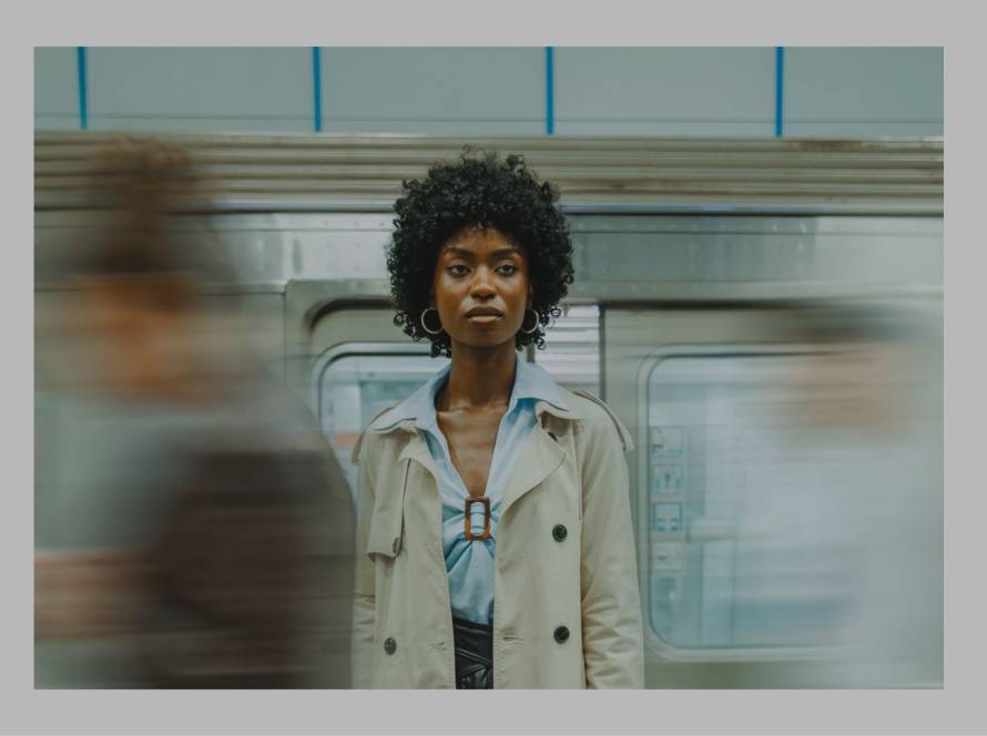 Woman standing composed on a train platform, motion blurred around her, embodying stillness amid surrounding movement