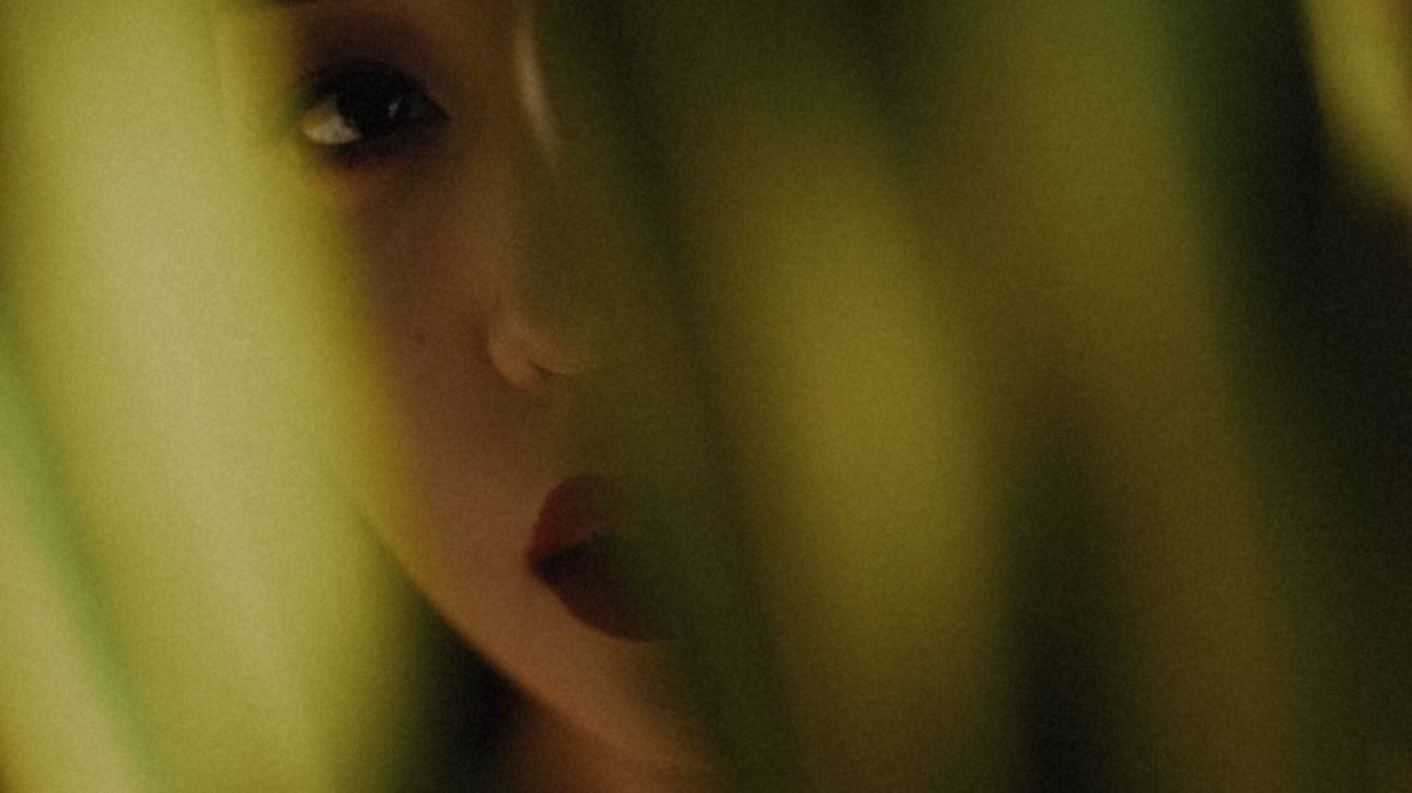 Close-up of a woman’s face, half hidden behind blurred green leaves, with one eye and her lips visible.