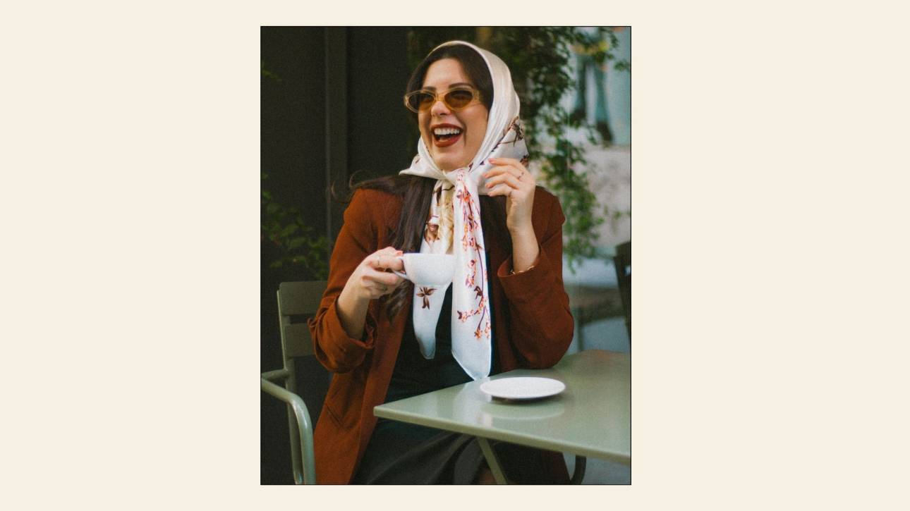Woman laughing at a café table, wearing tinted sunglasses, a cream floral headscarf and a rust jacket, holding a white coffee cup