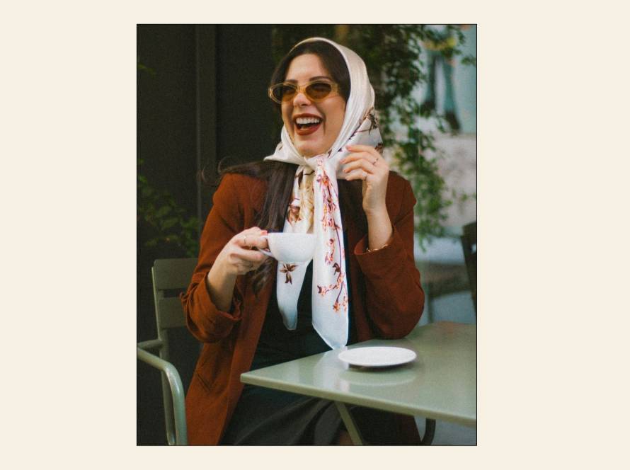 Woman laughing at a café table, wearing tinted sunglasses, a cream floral headscarf and a rust jacket, holding a white coffee cup